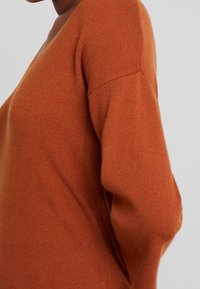 Maglione in maglia color ruggine con scollo a V, dotato di maniche lunghe e una consistenza morbida. Il tessuto presenta un sottile motivo a coste lungo le maniche.
