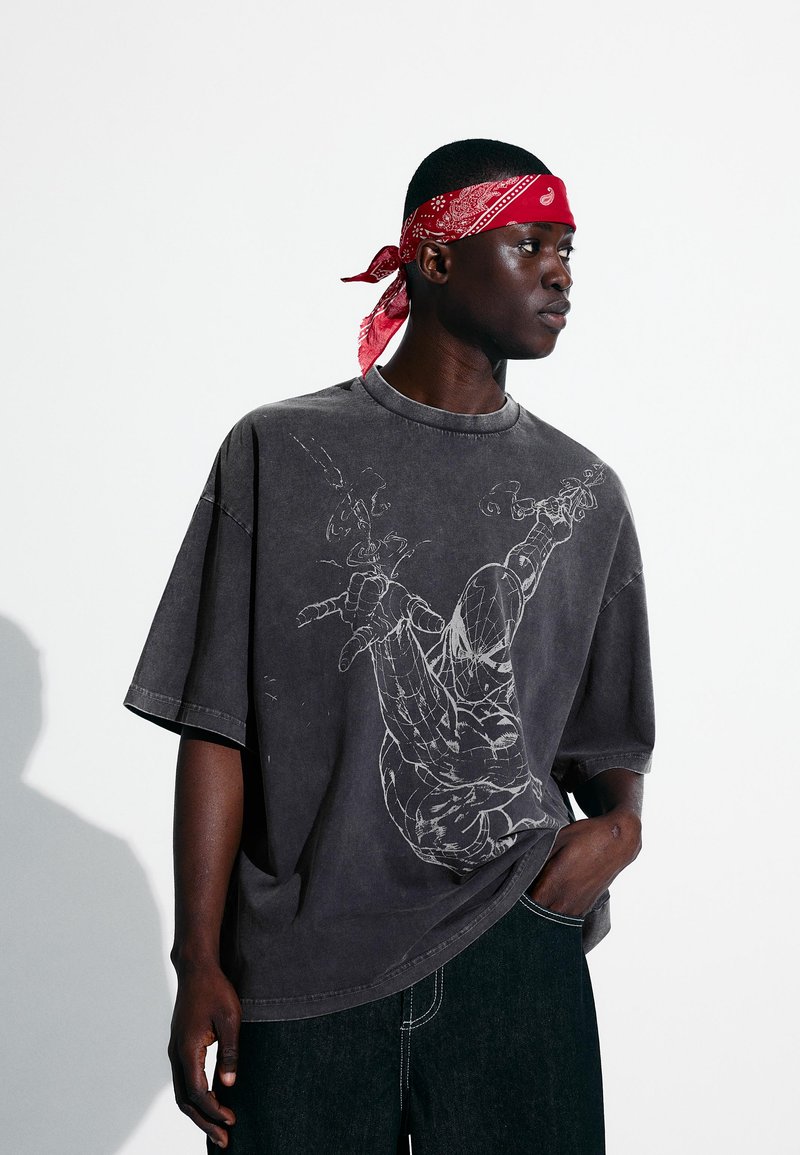 Mann trägt ein ausgebleichtes schwarzes Spider-Man-Grafik-T-Shirt, dunkle Jeans und ein rotes Bandana, steht mit einer Hand in der Hosentasche.