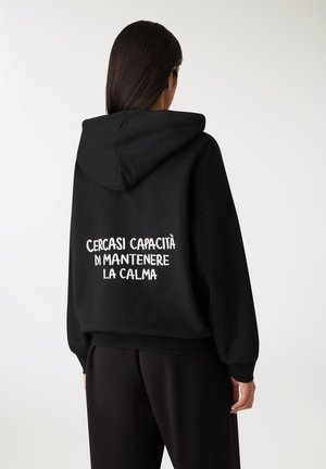 Felpa nera con cappuccio e vestibilità rilassata. Sul retro è presente un testo bianco: "CERCASI CAPACITÀ DI MANTENERE LA CALMA." Texture morbida.