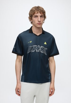 Junger Mann mit hellem lockigem Haar, der ein dunkelblaues ARKK-Sportshirt und hellfarbene Hosen trägt, steht vor einem schlichten weißen Hintergrund.