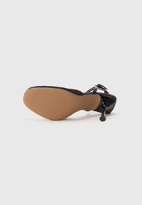 Scarpa nera con tacco alto in pelle lucida, cinturino alla caviglia regolabile e suola in gomma beige con punta leggermente appuntita.