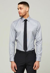 Ljusgrå skjorta med en subtil randi, långa ärmar, knappad krage, ihop med en svart slips och mörka byxor.