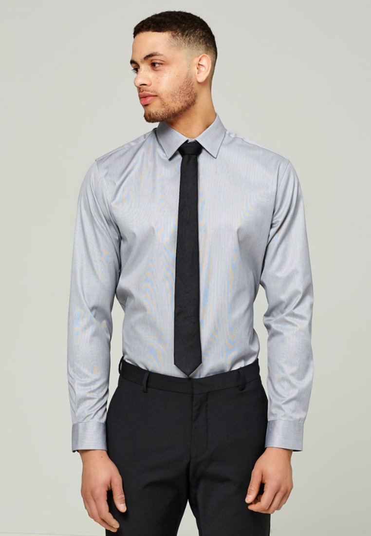 Ljusgrå skjorta med en subtil randi, långa ärmar, knappad krage, ihop med en svart slips och mörka byxor.