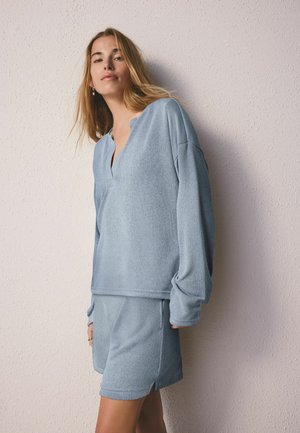 Fiatal nő hosszú szőke hajjal, világoskék kötött hosszú ujjú felsőt és hozzá illő rövidnadrágot visel, egy texturált, világos fal előtt áll.