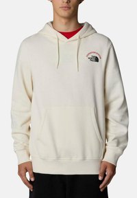 Sudadera beige con un bolsillo frontal, cordones y puños de canalé. Presenta un pequeño logo rojo y negro en el pecho. Textura suave.