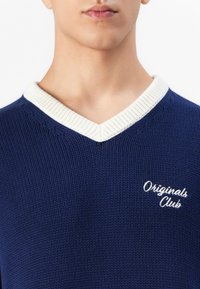 Gros plan sur une personne portant un pull en maille bleu en V avec des bordures blanches et "Originals Club" brodé sur la poitrine gauche.