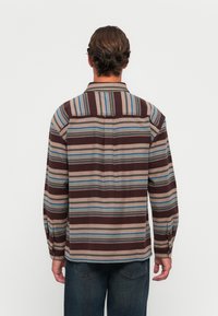 Man met kort bruin haar, gekleed in een lange mouwen gestreept shirt in bruin, beige, blauw en olijfgroene tinten, staand met zijn rug naar de kijker.