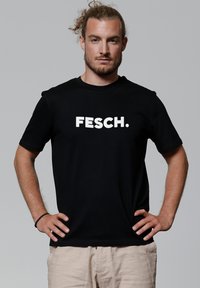 Zwarte katoenen t-shirt met korte mouwen, met daarop vetgedrukte witte tekst "FESCH." Vooraanzicht, model staat met handen op de heupen.
