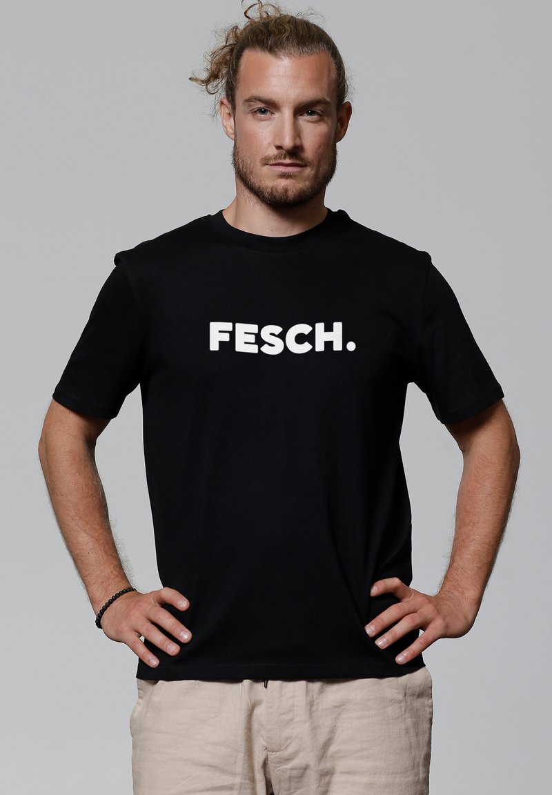 Zwarte katoenen t-shirt met korte mouwen, met daarop vetgedrukte witte tekst "FESCH." Vooraanzicht, model staat met handen op de heupen.