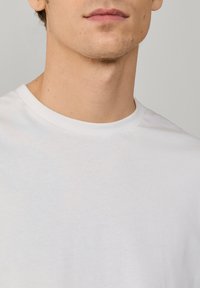 Junger Mann trägt ein schlichtes weißes T-Shirt mit Rundhalsausschnitt, zeigt unteren Teil des Gesichts, Hals und oberen Brustbereich vor neutralem Hintergrund.