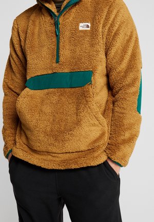 Brauner Fleece-Pullover mit türkisfarbenem Reißverschluss und Taschenklappe, getragen mit schwarzen Hosen, Hände in den Taschen, Oberkörper und Arme sichtbar.
