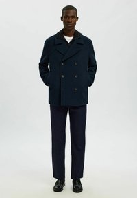 Cappotto peacoat doppiopetto blu navy con grandi bottoni neri e due tasche laterali, abbinato a pantaloni scuri e scarpe nere.