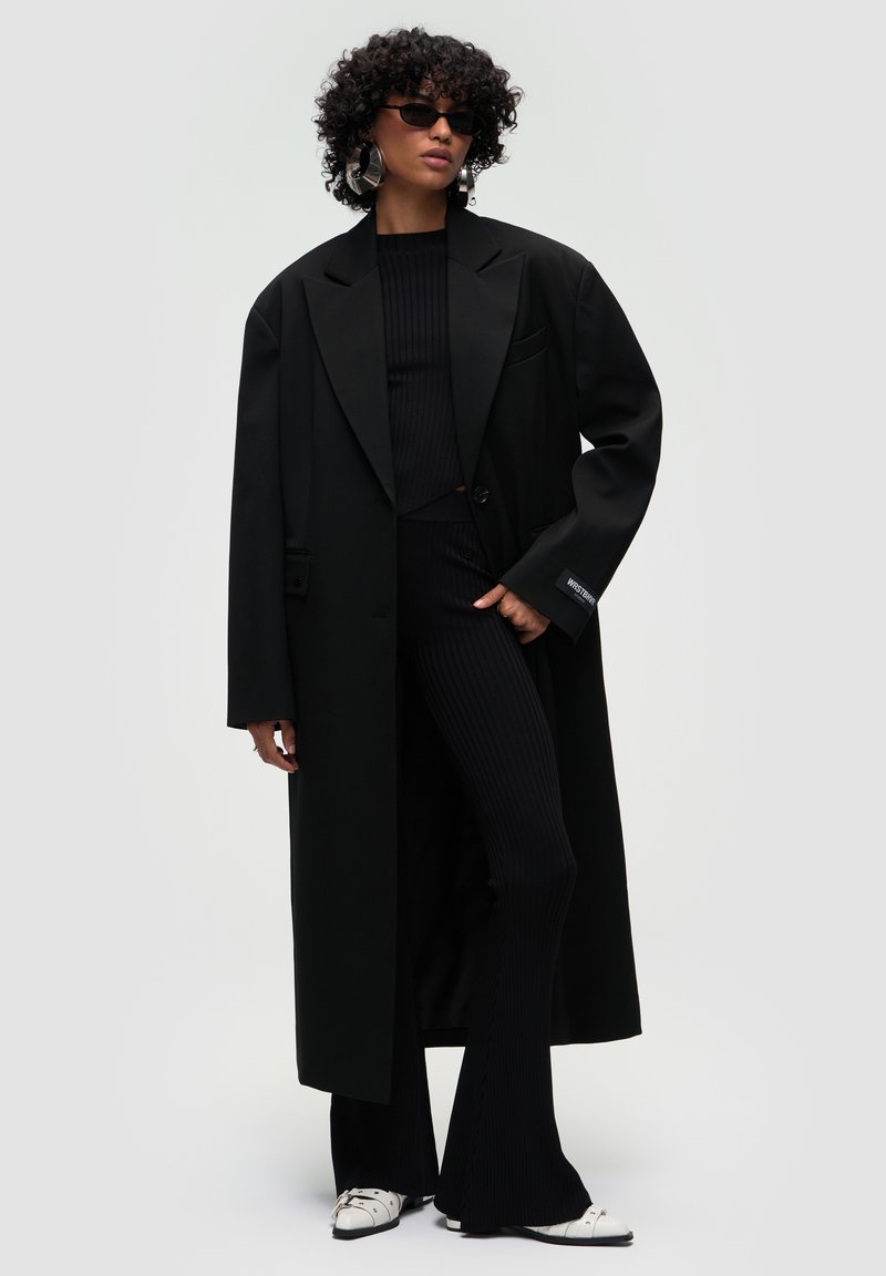 Cappotto nero oversize con colletto a punta, top nero a coste, pantaloni neri svasati e calzature moderne bianche. Gli accessori includono occhiali da sole e grandi orecchini.