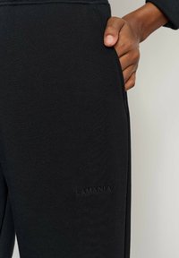 Pantaloni jogging negri din material moale, cu buzunar lateral și logo-ul „LAMANIA” în relief lângă talie. Textură netedă și croială mulată.