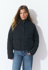 Veste noire matelassée avec un col haut, deux poches avant et un design court. La texture présente un motif géométrique.