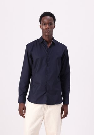Camisa de botonadura larga de color navy con cuello, que presenta una textura suave y botones oscuros. Llevada con pantalones de color claro.