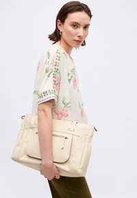 Γυναίκα με κοντά καστανά μαλλιά, που φοράει μπλούζα με λουλούδια και πράσινο παντελόνι, κρατώντας μια μεγάλη κρεμ τσάντα χειρός σε ένα απλό λευκό φόντο.