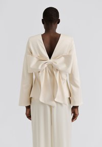 Kremfarget bluse med dyp V-rygg, som har en stor sløyfe og flytende folder ved midjen. Glatt tekstur og lange ermer.