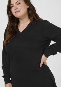 Nasmejana ženska z dolgimi rjavimi lasmi, oblečena v črno dolgotrajno srajco z V-izrezom, pozira z roko na boku.