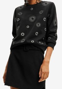 Czarny sweatshirt z wirującymi wzorami w srebrnej haftowanej formie, z okrągłym dekoltem i ściągaczami, zestawiony z czarną spódnicą i paskiem.