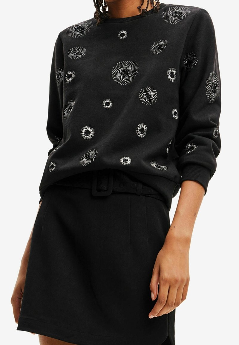 Czarny sweatshirt z wirującymi wzorami w srebrnej haftowanej formie, z okrągłym dekoltem i ściągaczami, zestawiony z czarną spódnicą i paskiem.