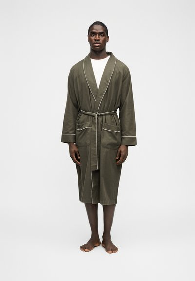 Άνδρας ξυπόλυτος, φοράει πράσινο χακί ρόμπα με ζώνη και λευκή επένδυση, καθώς και λευκό πουκάμισο από μέσα, πάνω σε απλό ανοιχτόχρωμο φόντο.