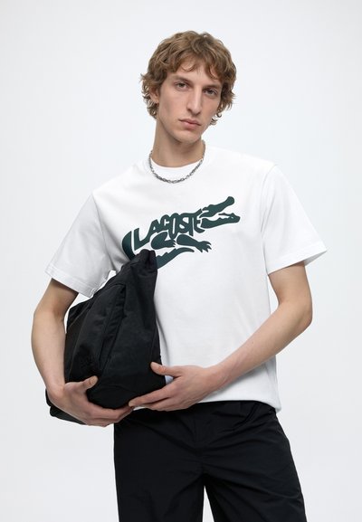 Млад мъж с вълниста коса, носещ бяла тениска Lacoste, държи черна раница на бял фон.