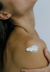 White cream is applied to a tanned shoulder. The skin is smooth, showcasing a natural glow, with a subtle texture and minimal detail around.
