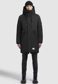 Melnā izolētā parka ar augstu apkakli, regulējamu kapuci, priekšējo rāvējslēdzēju, divām sānu kabatām un logo uzgabalu apakšmalā.