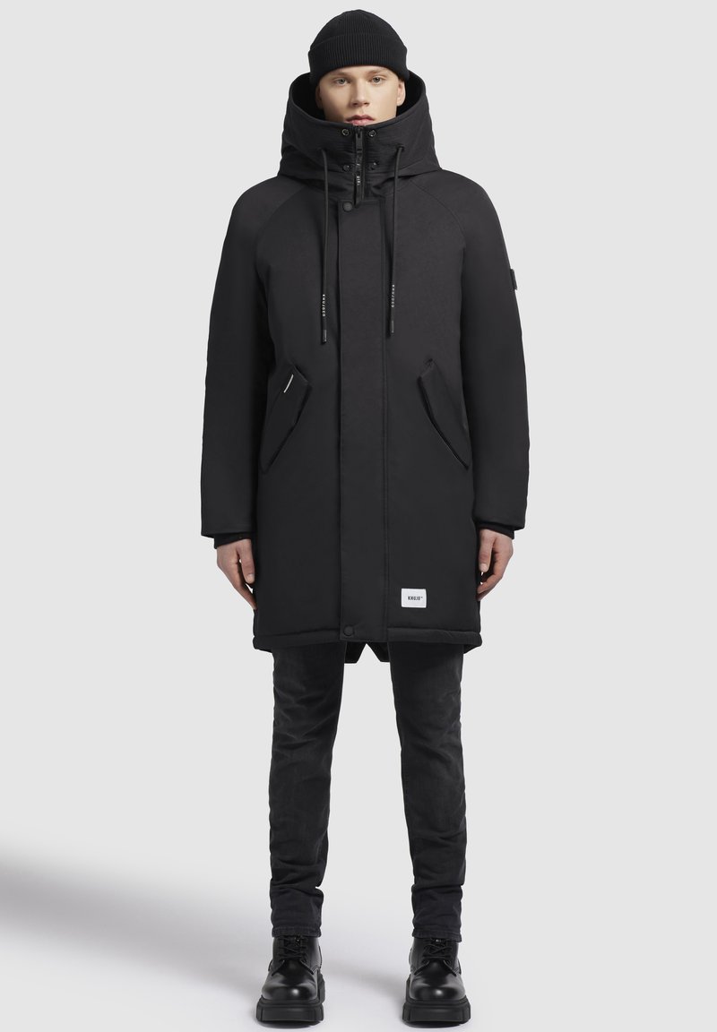 Melnā izolētā parka ar augstu apkakli, regulējamu kapuci, priekšējo rāvējslēdzēju, divām sānu kabatām un logo uzgabalu apakšmalā.