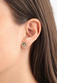 Pendientes de stud en tono dorado con un diseño circular, que presenta un detalle de estallido central sobre un fondo gris.