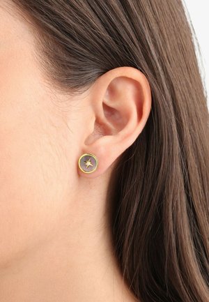 Pendientes de stud en tono dorado con un diseño circular, que presenta un detalle de estallido central sobre un fondo gris.