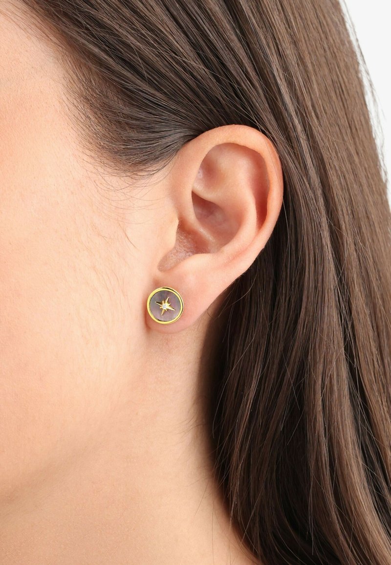 Pendientes de stud en tono dorado con un diseño circular, que presenta un detalle de estallido central sobre un fondo gris.