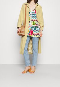 Beige trenchcoat, livlig mönstrad topp med blommor och zigzag-motiv, ljusblå jeans och gula klacksandaler. Vevad väska med brun rem.