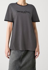 La camiseta gris de manga corta presenta un estampado gráfico negro. Tiene un cuello redondo con un ribete negro contrastante y un ajuste relajado.
