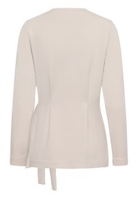 Blazer beige à manches longues avec coutures ajustées et deux liens en tissu pendant au bas du dos.