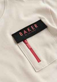 Kerman värinen t-paita, jossa on musta suorakulmainen kangas, jossa on punaista tekstiä. Edessä punaisella korostettuna vetoketjullinen tasku.