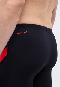 Κοντινό πλάνο ενός ατόμου που φοράει μαύρο μαγιό με κόκκινες πλευρικές λωρίδες και τη λέξη "ENDURANCE+" τυπωμένη με κόκκινα γράμματα στη μέση στην πίσω πλευρά.
