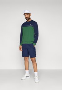 Uomo con cappello bianco, camicia a maniche lunghe verde e blu navy, pantaloni corti blu navy, calzini bianchi e scarpe da ginnastica bianche in piedi su uno sfondo neutro.