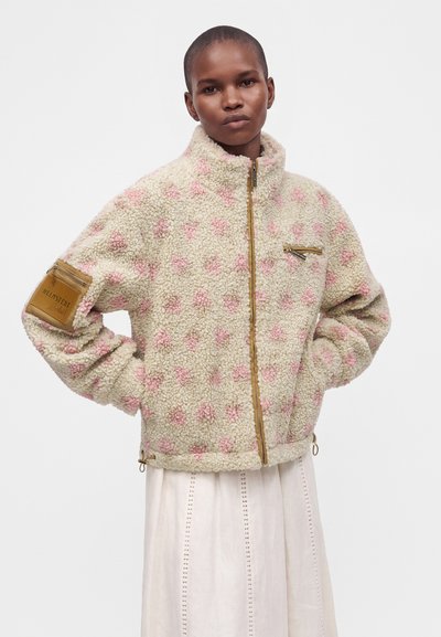 Personne aux cheveux courts portant une veste duveteuse beige et rose à motifs avec des poches zippées sur une jupe blanche.