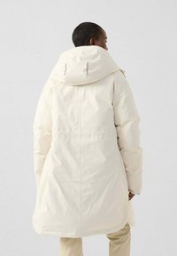 Persona de espaldas vistiendo un largo abrigo acolchado de invierno en un tono blanco roto con un gran capucho y pantalones beige, contra un fondo sencillo.