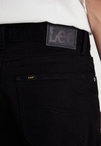 Schwarzer Lee-Jeansbund und Detail der Gesäßtasche mit sichtbaren Markenetiketten, getragen mit einem weißen Oberteil.