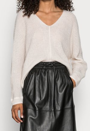 Femme portant un pull blanc en maille à col en V rentré dans une jupe en cuir noire taille haute avec poches et taille à cordon.