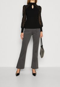 Pull noir avec des manches bouffantes en dentelle et un décolleté en trou de serrure, associé à un pantalon gris évasé et des escarpins noirs à bout pointu, tenant un sac en maille.