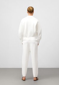 Sweatshirt blanc et pantalon assorti en tissu doux et texturé ; coupe décontractée avec des poignets côtelés et un design simple. Tongs noires visibles.
