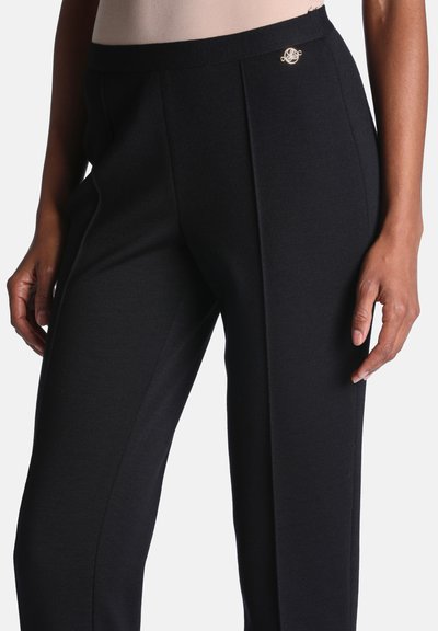 Femme portant un pantalon noir cintré avec un pli devant et un petit emblème rond doré près de la taille côté droit.