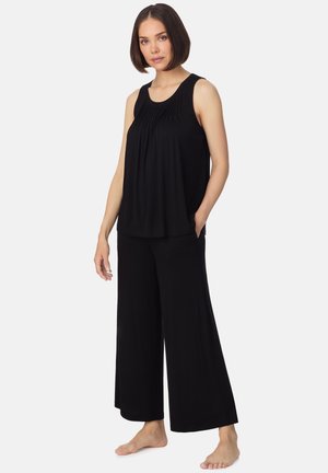 Kobieta o krótkich brązowych włosach, ubrana w czarny plisowany top bez rękawów i szerokie czarne spodnie, stojąca boso na jednolitym tle.