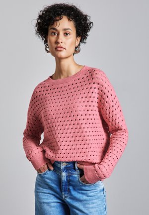 Locker gestrickter rosa Pullover mit runden Lochmustern, weitem Rundhalsausschnitt und langen Ärmeln. Kombiniert mit hellblauen Denim-Jeans.