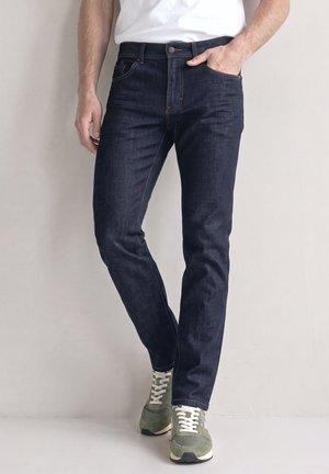 Uomo con jeans blu scuro e sneaker verdi, in piedi con una mano in tasca davanti a uno sfondo uniforme.