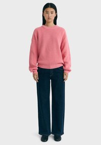 Un pull rose à côtes avec des manches longues, associé à un jean large et foncé. La tenue présente une coupe décontractée et une palette de couleurs unie.